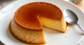 Un flan de dulce de leche con caramelo brillante en la parte superior, una porción ya cortada y una cuchara de metal a su lado, servido en un plato blanco.