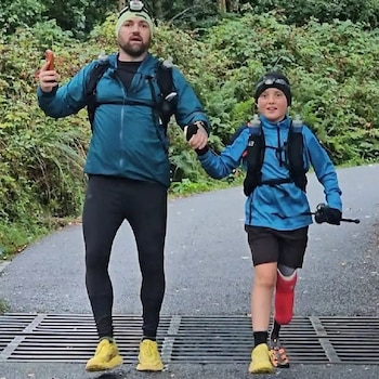 Un hombre y un niño con una prótesis roja en la pierna izquierda caminan tomados de la mano sobre una rejilla metálica en un camino