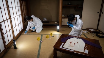 Dos técnicos forenses trabajan en una vivienda japonesa con suelo de tatami, entre tres marcadores de evidencia, luces y una remera con sangre.