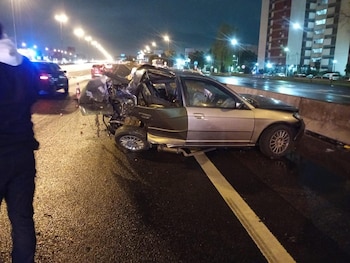 Un automóvil gris con la parte trasera destruida se encuentra sobre el asfalto mojado de una vía urbana iluminada por farolas