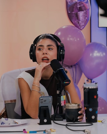 Cami Mayan sonríe sosteniendo un ramo de flores envuelto en papel blanco, vestida con top oscuro, jeans y cinturón dorado, frente a unas escaleras de madera