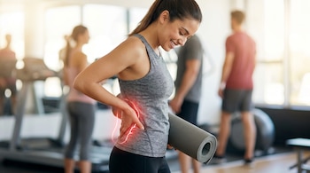 Mujer joven de pie en un gimnasio, sonriendo y tocándose la espalda baja con una mano, mientras sostiene un tapete de yoga enrollado. La zona lumbar está marcada en rojo.
