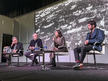 Cuatro personas sentadas en un escenario oscuro; la mujer central habla por micrófono. Detrás, una pantalla grande proyecta una imagen granulada en blanco y negro
