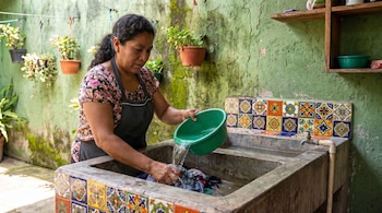 Mujer de blusa floral y delantal vierte agua de balde verde en lavadero de piedra con ropa. Pared verde con plantas y estantes.