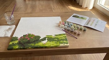 Un lienzo de pintura por números a medio pintar sobre una mesa de madera con tarros de pintura, pinceles, un vaso con agua y un manual.