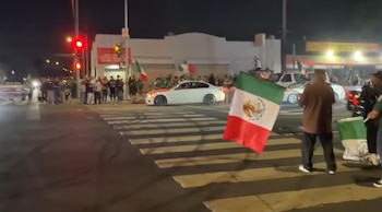 Vista nocturna de una calle con personas y varios coches, algunos con banderas de México, y un semáforo en rojo. Huellas de neumáticos en el asfalto