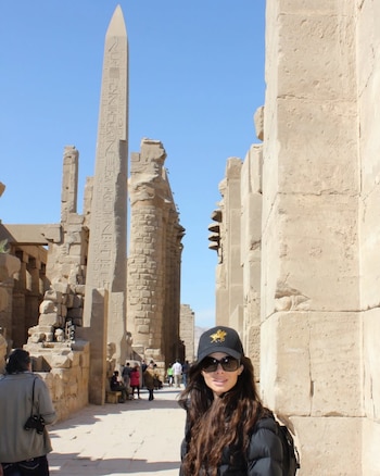 Mujer sonriente con gorra negra y gafas de sol frente a un alto obelisco de piedra con jeroglíficos y columnas de piedra antiguas bajo un cielo azul claro