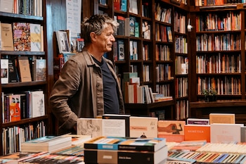 Un hombre con chaqueta marrón está de pie frente a estanterías de madera repletas de libros en una librería.