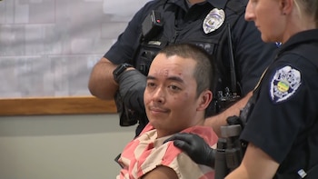 Un hombre con uniforme a rayas y marcas en el rostro permanece sentado mientras dos oficiales de policía con guantes negros lo sostienen