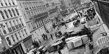 Vista aérea en blanco y negro de una calle de la ciudad con numerosos coches volcados y personas, formando barricadas entre edificios