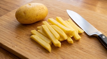 Una patata entera, bastones de patata pelada y cortada, y un cuchillo de chef con mango negro sobre una tabla de madera.