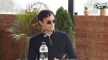 Un hombre con gafas de sol y suéter azul habla sentado frente a una botella de agua, con plantas y una pared de ladrillos al fondo