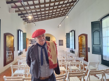 Un hombre con boina roja y una manta sobre el hombro está de pie ante filas de sillas blancas en una sala con techo de madera