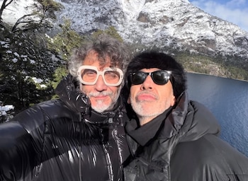 Dos hombres se toman una selfie frente a un lago, montañas nevadas y árboles. Uno lleva gafas de marco blanco, el otro gafas de sol y gorro de piel negro
