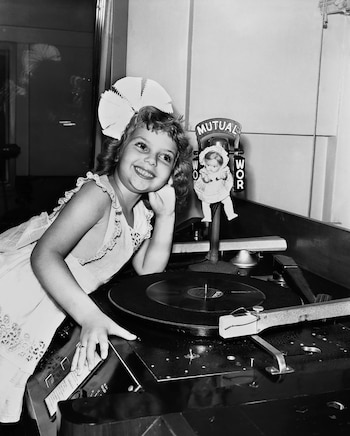 Niña sonriente con un lazo grande en el cabello posa apoyada en un equipo tocadiscos junto a un micrófono antiguo