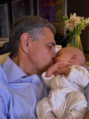 Primer plano de un hombre canoso con camisa azul besando la cabeza de un bebé durmiendo, vestido con un mameluco blanco