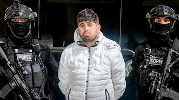 Un hombre con chaqueta blanca permanece custodiado por dos policías fuertemente armados con uniforme táctico negro y pasamontañas