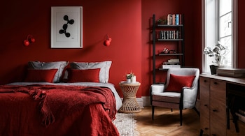 Una vista completa de un dormitorio con paredes rojas, una cama grande con ropa de cama roja y gris, una estantería, un sillón y una mesita lateral.