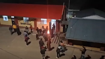 Vista de noche desde una altura de un grupo de personas reunidas frente a un edificio con techo rojo y un fuego pequeño en la entrada