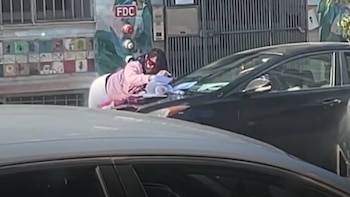 Una mujer con blusa rosa se inclina sobre el capó de un sedán deportivo oscuro, pareciendo limpiar algo. Parte de otro coche claro está en primer plano