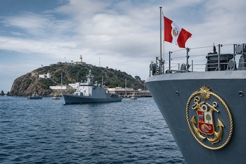 Proa de buque náutico con la bandera peruana y el escudo de la Marina, con otra embarcación y la isla de San Lorenzo al fondo.