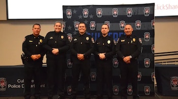 Cinco agentes de policía uniformados están de pie frente a un panel oscuro con logotipos institucionales y una mesa cubierta