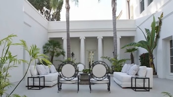 Patio exterior blanco con dos sofás, dos sillas de metal negro, una mesa y plantas tropicales. Edificio con columnas al fondo