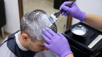 Manos con guantes morados aplican una mezcla blanca con una brocha sobre el cabello corto de un hombre en una peluquería.