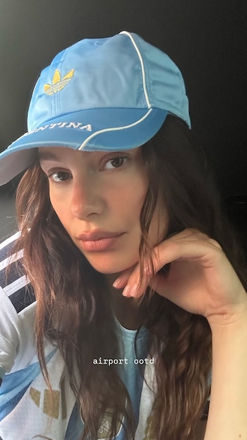 Una mujer con cabello oscuro y gorra celeste con el logo de Adidas y la palabra Argentina. Viste una camiseta a rayas celestes y blancas