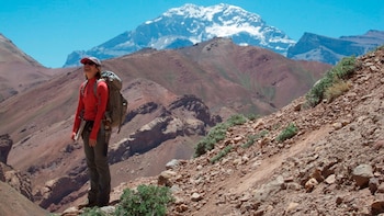 Científica Barbara Carrapa en una ladera rocosa, vestida de rojo, con mochila, observando montañas áridas y un pico nevado bajo un cielo azul