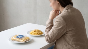 Persona sentada en una mesa blanca con un plato de pasta cocida y un paquete de pasta sin gluten con etiqueta azul.