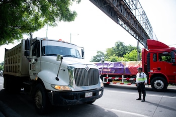 Un camión blanco de volteo a la izquierda, un camión rojo con carga cubierta a la derecha y un agente uniformado en la calle