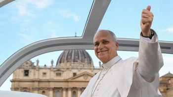 Un hombre con sotana blanca levanta el pulgar dentro de un vehículo transparente con una cúpula y una basílica al fondo