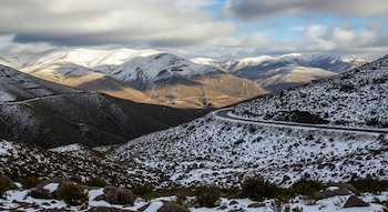 Vista de un paisaje serrano invernal con montañas parcialmente nevadas y una carretera sinuosa. El sol ilumina algunas laderas bajo un cielo nublado.