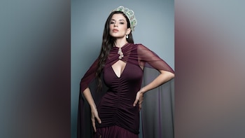 Mujer de cabello oscuro con vestido color vino, corona de joyas verdes y transparentes, y pendientes de perlas posa con manos en caderas