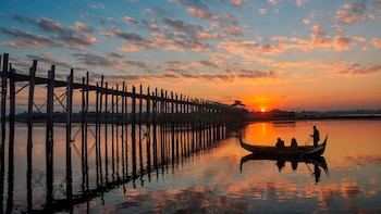 Un largo puente de madera sobre agua, un bote con tres personas a bordo, el sol visible en el horizonte, cielo con nubes azules y anaranjadas
