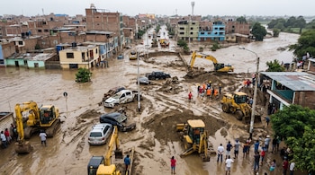 Calle urbana inundada con lodo, casas anegadas, varios coches dañados y maquinaria pesada, incluyendo excavadoras, que trabaja en la zona, mientras personas observan los daños.