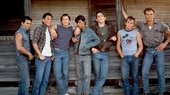 Siete jóvenes, incluyendo a Matt Dillon y Patrick Swayze, posan de pie contra una pared de madera rústica, vestidos con ropa informal de los años 80