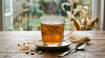 Taza de vidrio transparente con té humeante y hebras en un plato, sobre mesa de madera con granos de maíz y una cuchara.