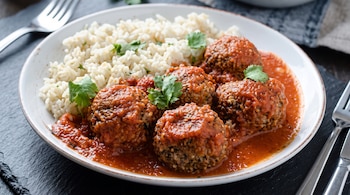 Un plato blanco con albóndigas en salsa de tomate, arroz blanco y cilantro, dispuesto sobre una superficie oscura junto a tenedores.