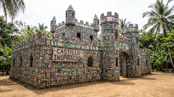 Un castillo edificado con botellas de plástico sobre un terreno de tierra, con un cartel de madera que dice Castillo Inspiración y palmeras.
