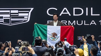 Checo Pérez sostiene la bandera de México frente a un panel de Cadillac Formula 1 mientras varias personas le toman fotografías.