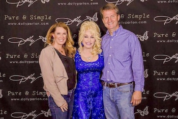 Dolly Parton, vestida de azul, sonríe entre una mujer y Tim Burchett frente a un fondo promocional de la cantante