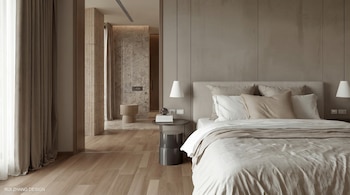 Un dormitorio moderno con una cama grande y sábanas blancas, almohadas de colores neutros, dos lámparas colgantes y mesas de noche. Un baño con paredes de piedra es visible al fondo.