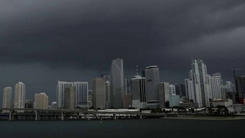 Ciudad costera con rascacielos y edificios altos bajo un cielo de tormenta oscuro. Se observan un cuerpo de agua en primer plano y un puente