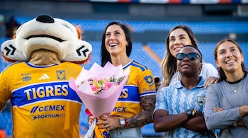 Un león mascota, una mujer con jersey de Tigres y un ramo de flores, y otras tres mujeres en un estadio de asientos azules.