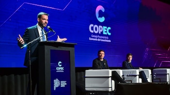 Hombre en traje habla por micrófono en atril, frente a pantalla azul con logo COPEC y texto, con dos hombres sentados en primera fila