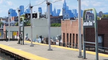 Plataforma de metro con franja amarilla y postes. Un letrero dice 'Junction Blvd'. Al fondo, edificios de ladrillo y un horizonte de rascacielos bajo un cielo azul