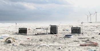 Tres amplificadores, una mesa con cables y dos micrófonos sobre la arena de una playa con basura y aerogeneradores al fondo