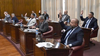 Varios legisladores de traje sentados en escritorios de madera dentro de un recinto legislativo levantan la mano derecha